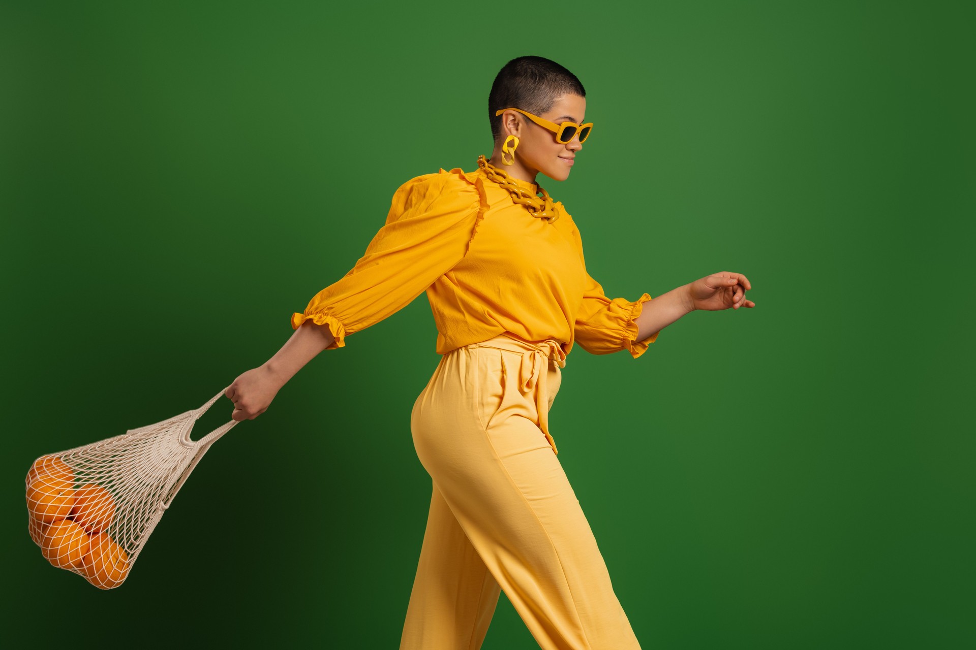 Beautiful young short hair woman carrying mesh bag with oranges against green background